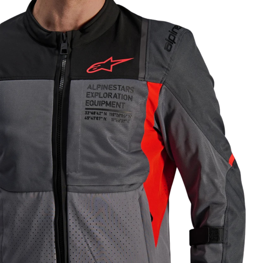 Close-up of a person wearing an Alpinestars technical jacket, showing the chest pocket with 'ALPINESTARS EXPLORATION EQUIPMENT' text, red logo, and bright red side panel accents on a dark gray and black base.