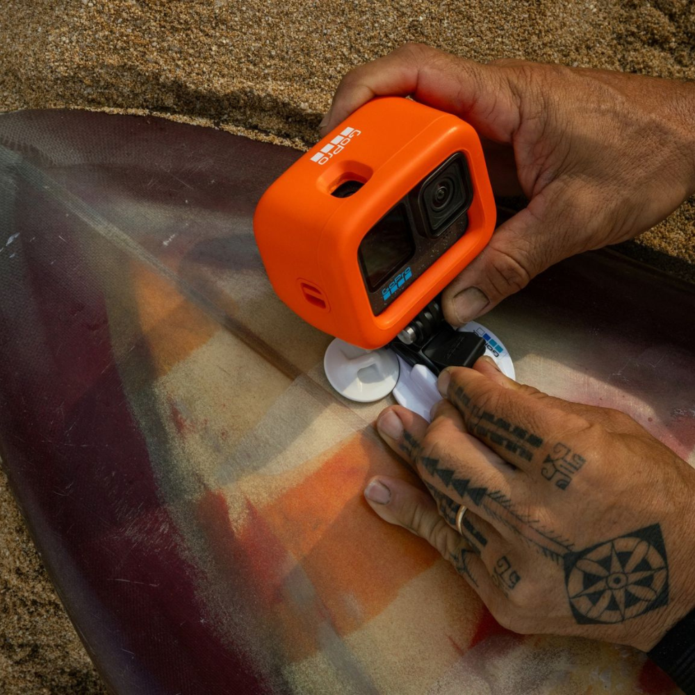 An orange protective case for a GoPro camera is shown mounted on a surfboard, with a person's tattooed hands adjusting the mounting hardware on a sandy beach.