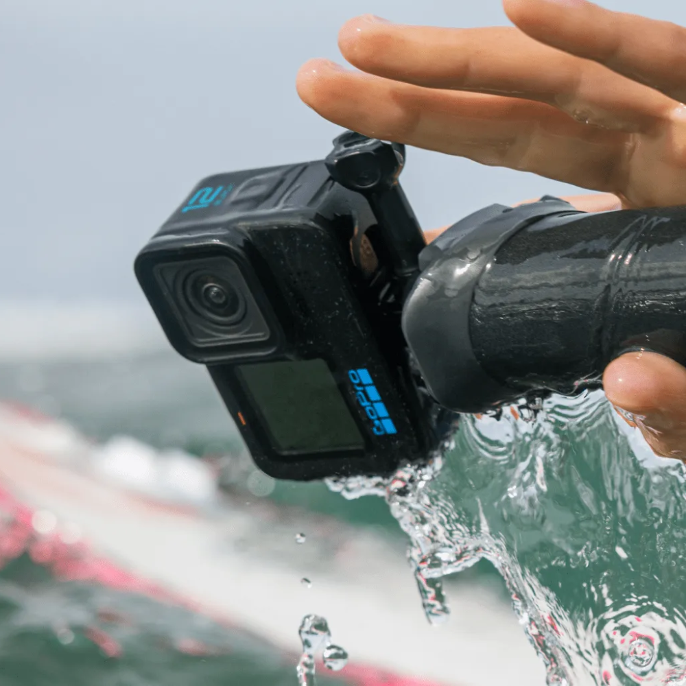 A black GoPro action camera with blue branding is shown being held underwater or under running water, with a hand visible above it and a blurred outdoor background.