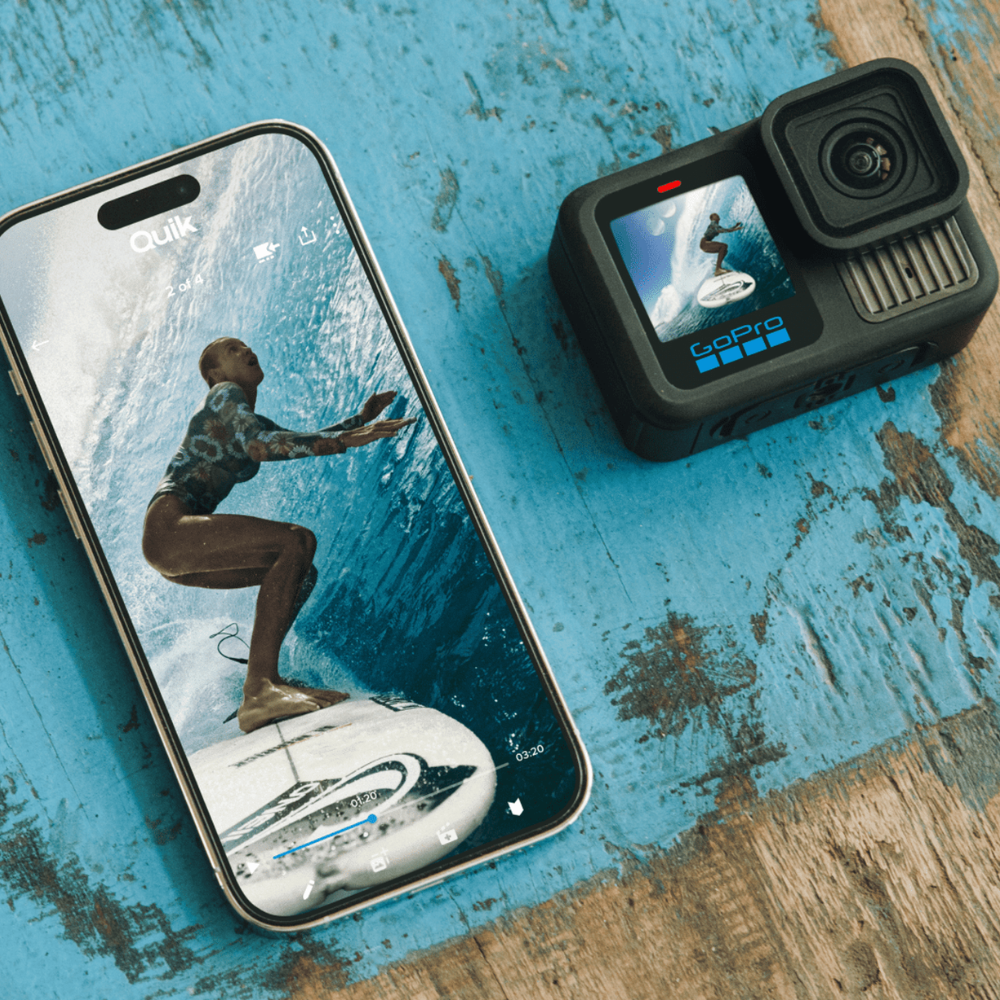 A GoPro action camera placed on a distressed blue wooden surface next to a smartphone displaying a surfing scene. The camera shows the GoPro logo on its screen bezel.
