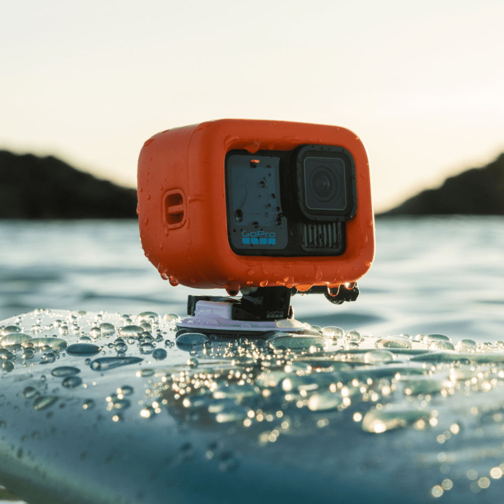 An orange waterproof housing for a GoPro action camera, shown mounted on a surface with water droplets, set against a blurred outdoor water and mountain background.