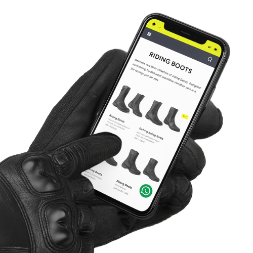 Hand wearing a black glove holding a smartphone displaying a riding boots product page.