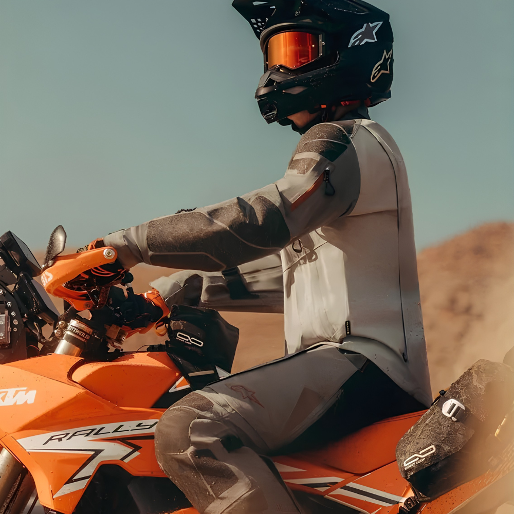 A rider wearing a black Alpinestars full-face helmet with orange-tinted goggles, a gray and black riding jacket, gloves, and matching pants, seated on an orange KTM motorcycle in a desert setting.