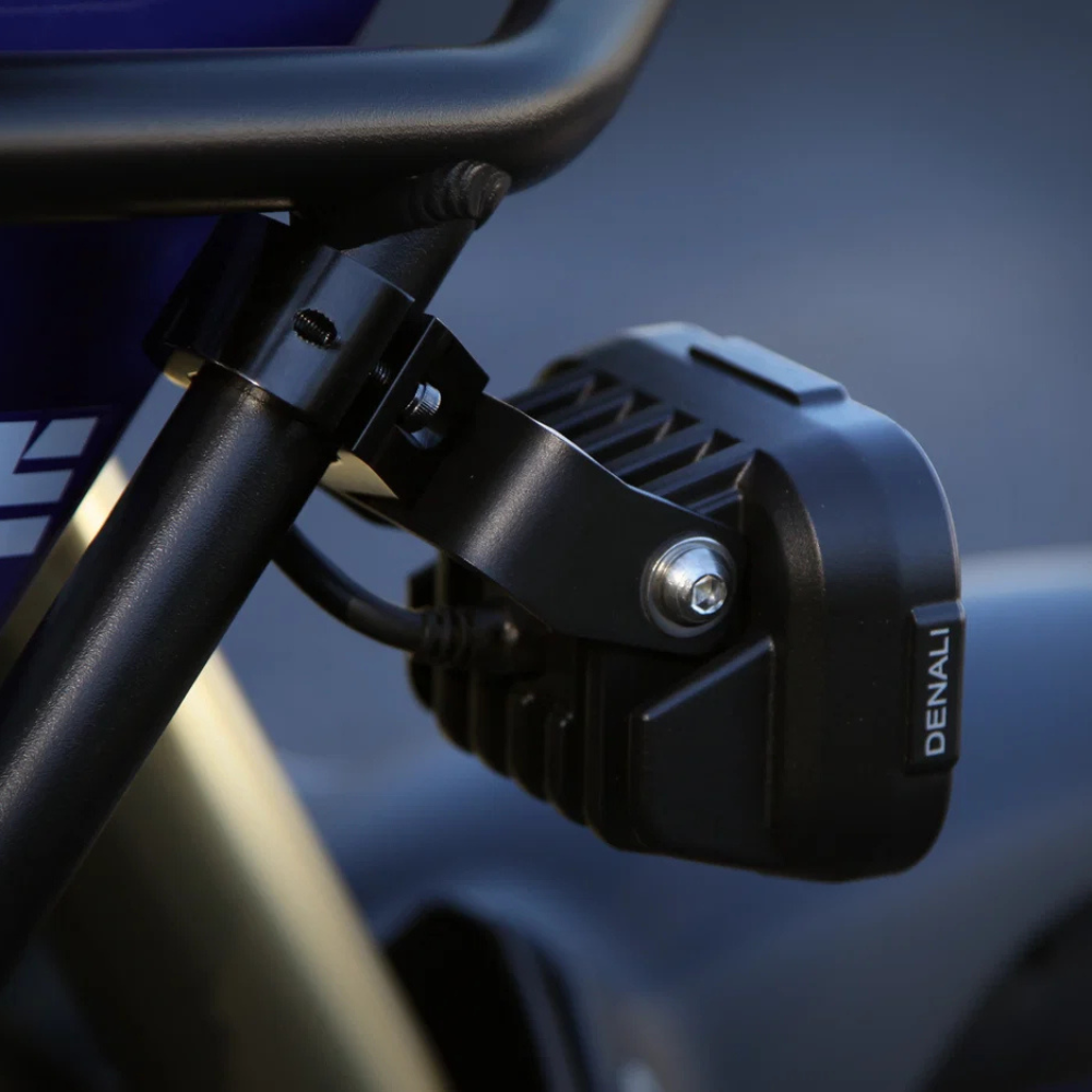 Close-up of a black Denali branded motorcycle or ATV headlight mounted on a black metal frame, with a blurred blue-gray background.
